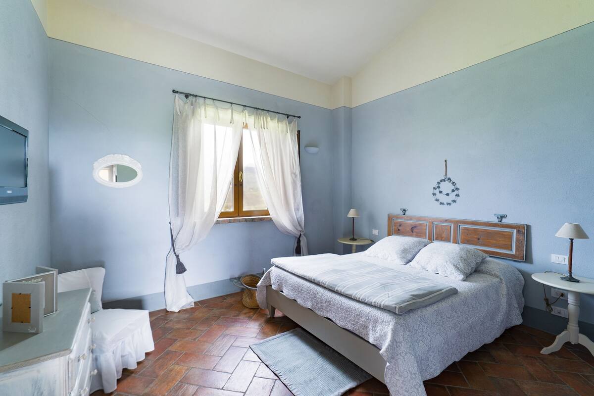 A bedroom with a bed, bedside tables, a mirror, a window with curtains, and a wall-mounted lamp.