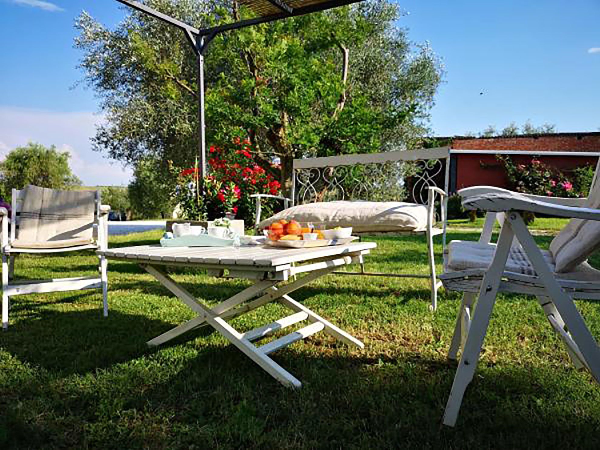 Un patio exterior con una mesa dispuesta para comer, rodeado de sillas, y un jardín con árboles y flores al fondo.