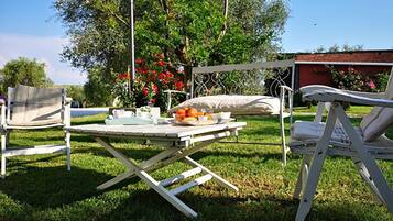 Un patio exterior con una mesa dispuesta para comer, rodeado de sillas, y un jardín con árboles y flores al fondo.