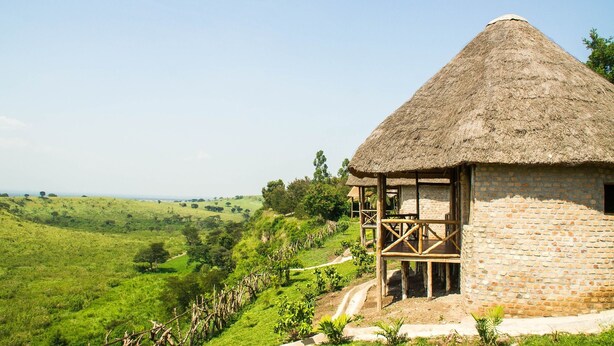 A thatched-roof hut with a wooden balcony, surrounded by lush green hills and a clear sky.
