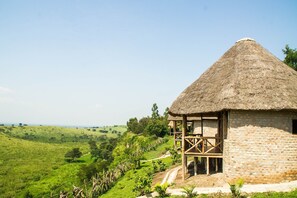 A thatched-roof hut with a wooden balcony, surrounded by lush green hills and a clear sky. - Elephant HAB Lodge (Kichwamba)