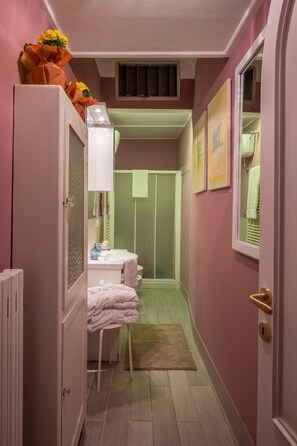 Un pasillo estrecho con un baño a la derecha, que incluye una ducha, un lavamanos y un inodoro. Hay toallas apiladas en una silla y una alfombra pequeña en el suelo.