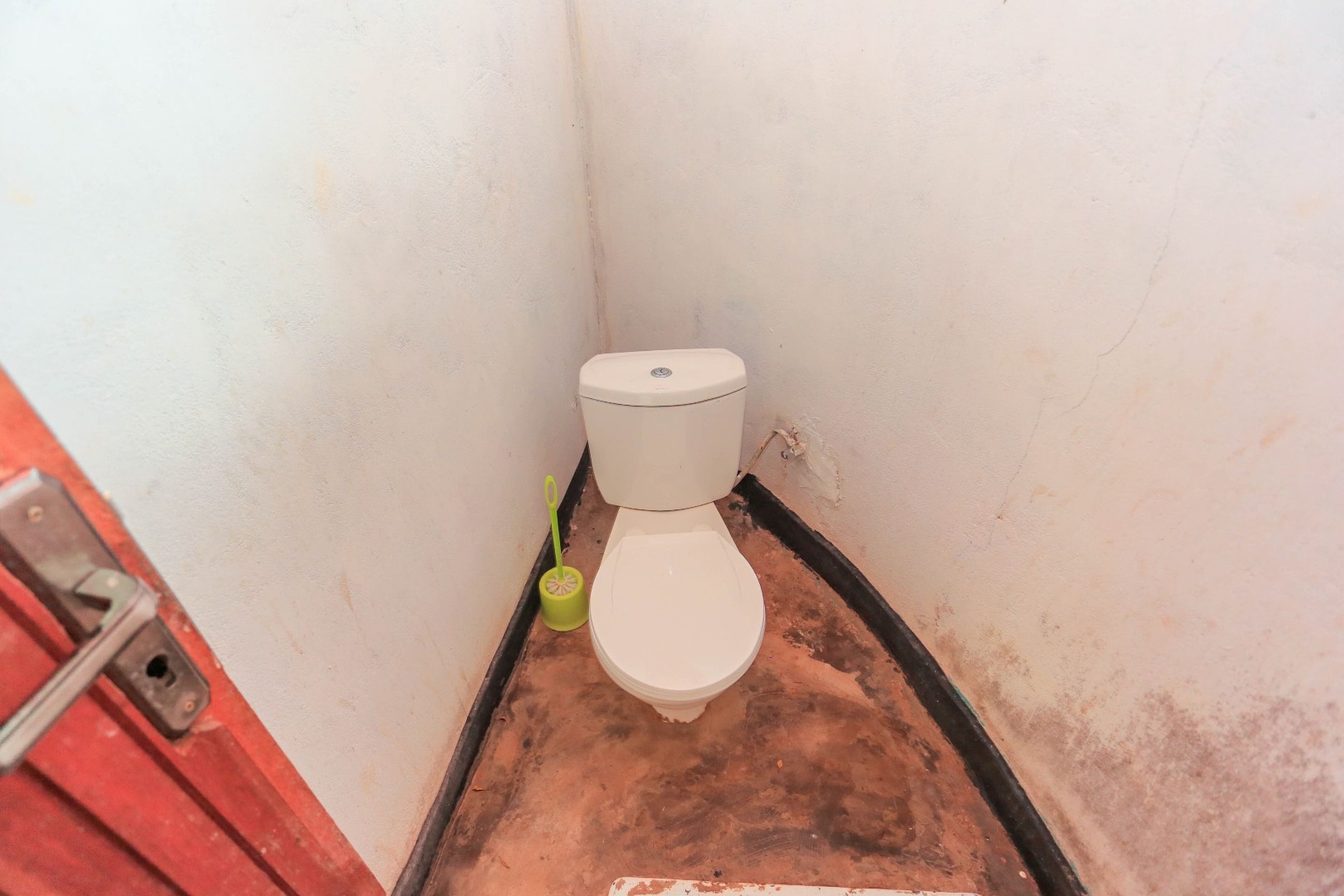 Un baño pequeño y estrecho con inodoro blanco, un cepillo verde de limpieza y una puerta roja.