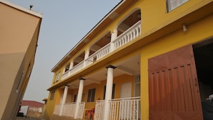 Un edificio amarillo con balcón, una puerta roja y una escalera que lleva a la entrada.