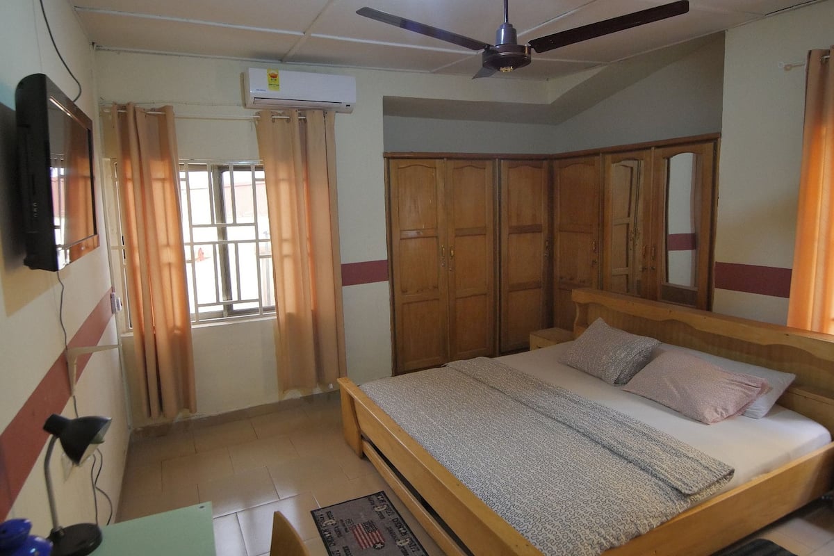 A bedroom with a bed, wooden wardrobe, ceiling fan, and a TV mounted on the wall.