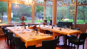 Een restaurant met meerdere tafels klaar voor diners, grote ramen met uitzicht op het omliggende landschap, en een kroonluchter aan het plafond.