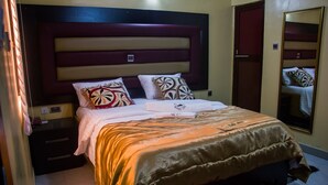 A neatly made bed with a patterned bedspread, a headboard with horizontal panels, and a bedside table with a lamp. - Sylva Link Suites (Lekki)