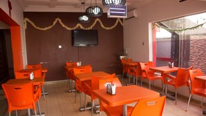 A dining area with orange tables and chairs, a flat-screen TV on the wall, and a view of the outside. - Sylva Link Suites (Lekki)