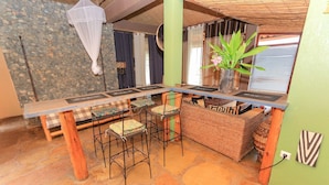 Een rustiek interieur met een stenen vloer, een lange houten tafel met barstoelen, een geweven bank en een hangend klamboe.