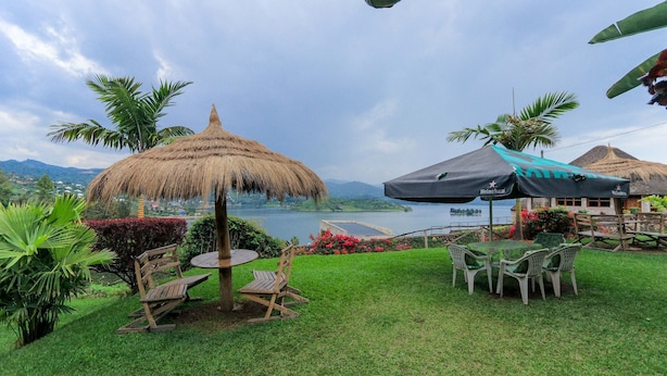 Een terras met een tafel en stoelen, een rieten parasol en uitzicht op een meer en bergen.