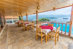 A covered terrace with wooden tables and chairs, overlooking a lake and mountains. - Nirvana Heights Hotel (Nyayumba)