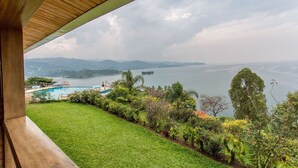 Vista desde un balcón sobre una piscina, exuberante vegetación y una masa de agua distante con montañas al fondo.