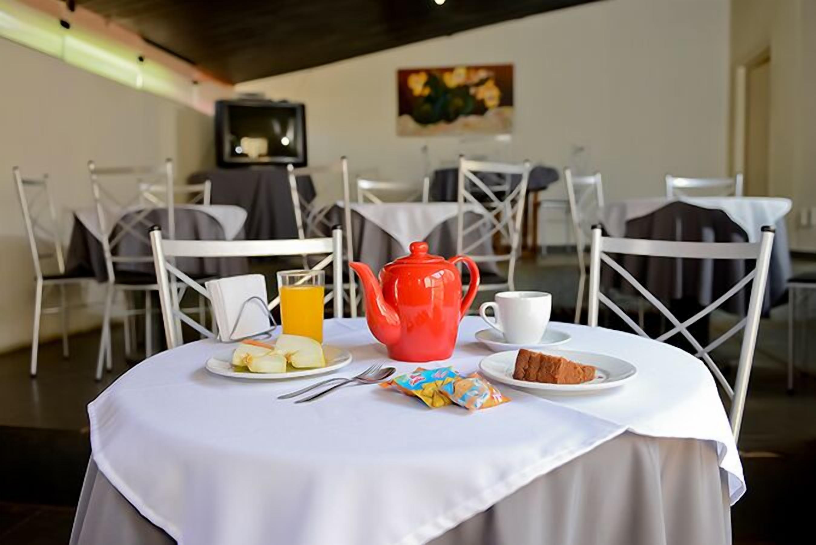 Mesa de café da manhã posta com uma chaleira vermelha, uma xícara branca, um copo de suco de laranja, um prato com frutas fatiadas, um pacote de lanches e uma fatia de bolo.