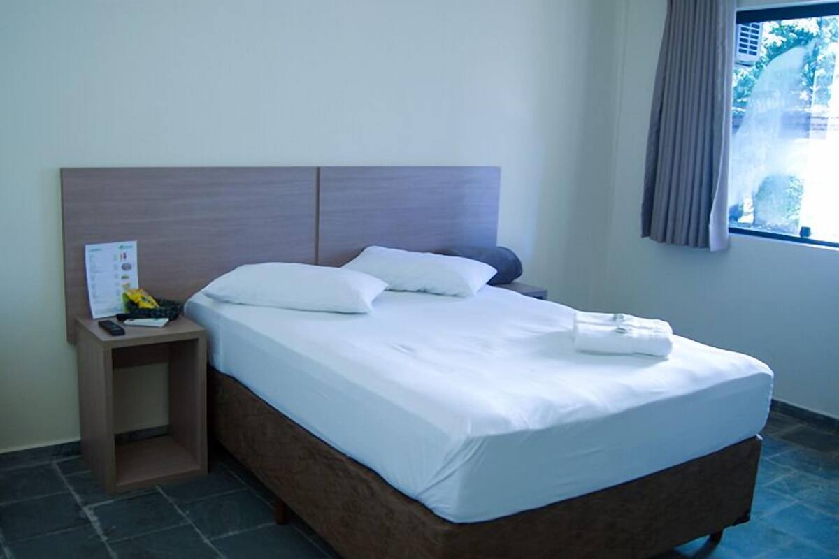 A hotel room with a bed, a nightstand, a small table, a window with curtains, and a view of greenery outside.