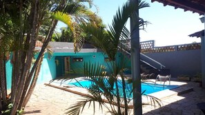Una piscina con solárium de baldosas, rodeada de palmeras y un edificio azul.