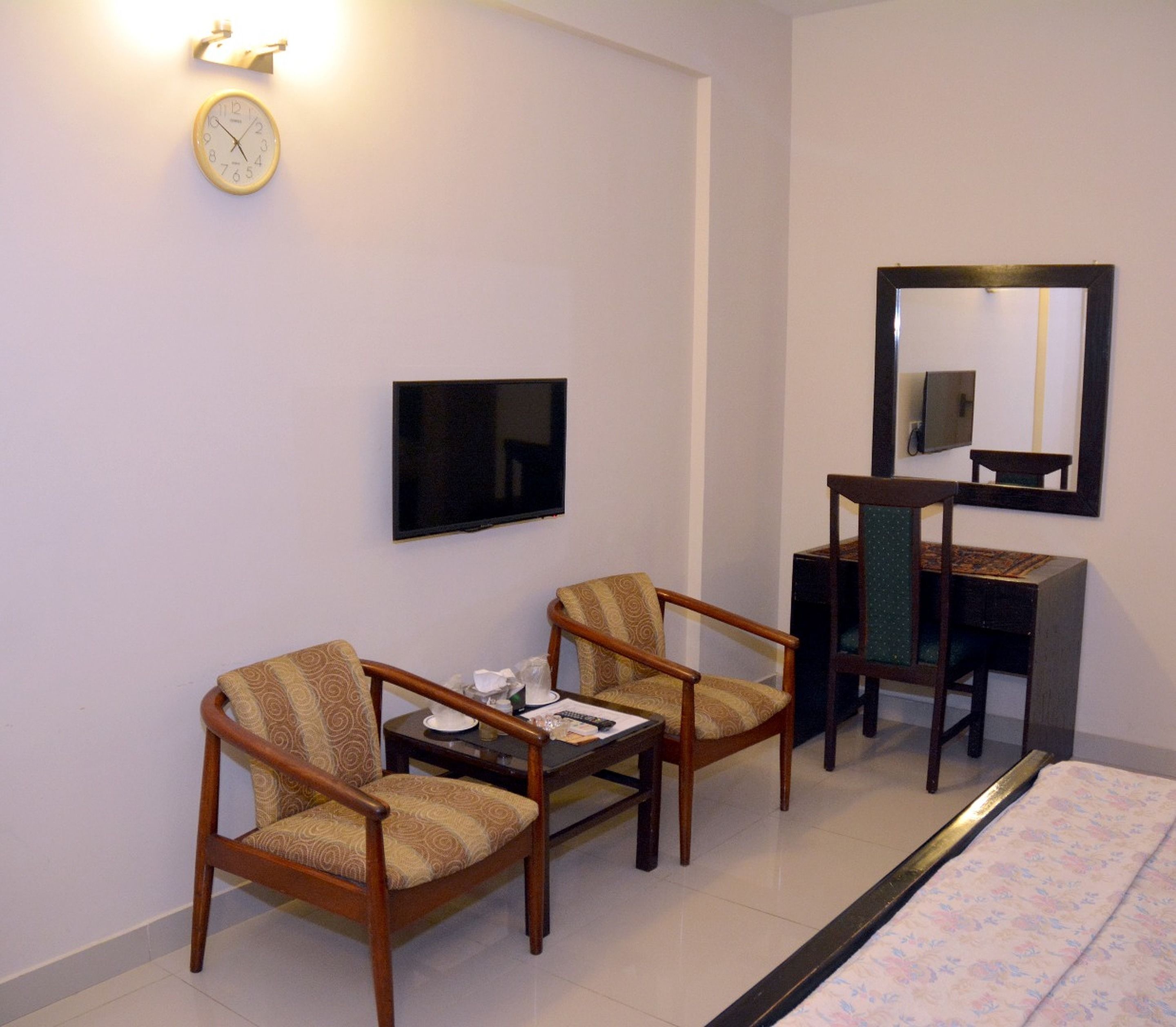 Habitación de hotel con dos sillones, una mesita, un escritorio con silla, televisor montado en la pared y un reloj.