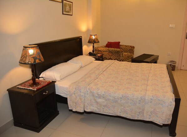 A neatly made bed with a floral bedspread, two bedside lamps, a nightstand, a small table with a red pillow, and a framed picture on the wall.