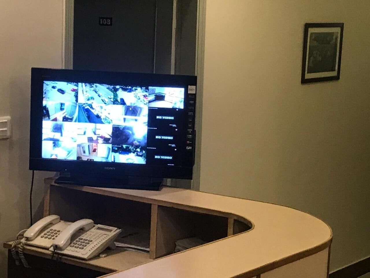 Zona de recepció d'hotel amb un televisor de pantalla plana que mostra diverses càmeres de vigilància, un telèfon i una porta amb el número 103.