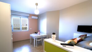 A small, well-lit room with a dining table, chairs, a white sofa, a TV mounted on the wall, and a window with a view of palm trees. - La Palma del Sol (Barletta)