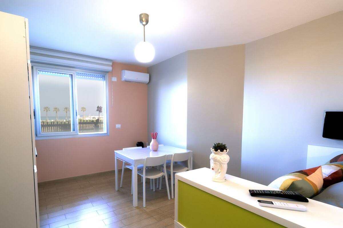Une petite pièce bien éclairée, avec une table à manger, des chaises, un canapé blanc, un téléviseur fixé au mur et une fenêtre donnant sur des palmiers.