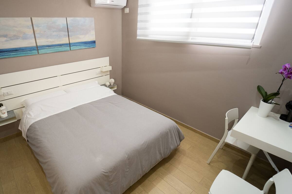 Une chambre à coucher avec un lit, un bureau, une chaise, une plante en pot et un tableau accroché au mur.