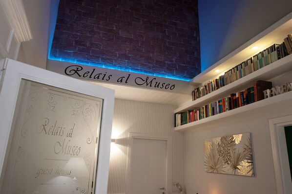 Huoneessa on kirjahylly, kehystetty kyltti, jossa lukee ”Relais al Museo -vierastalot”, ja ovi.