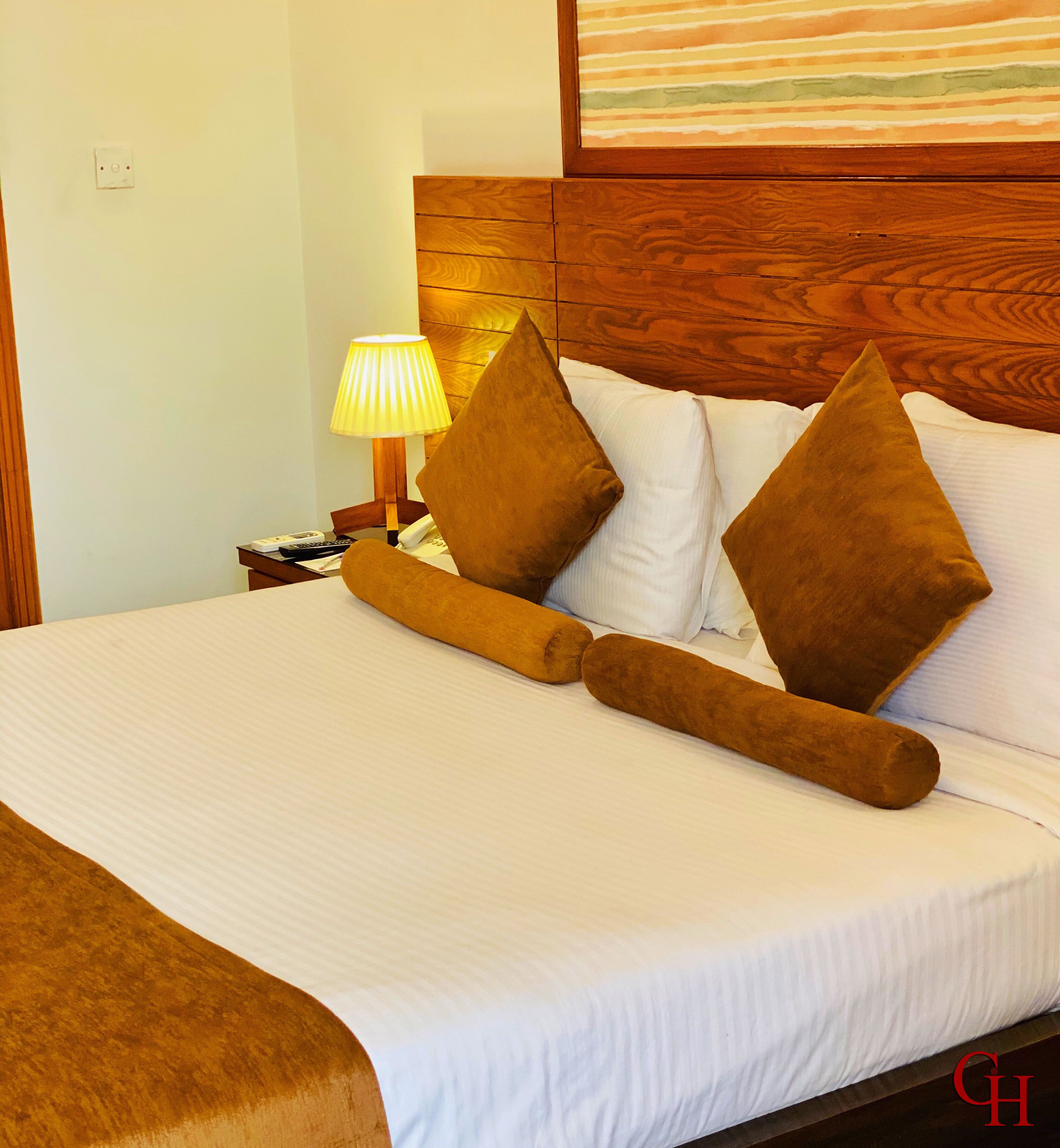 Una cama bien hecha con sábanas blancas y dos almohadas marrones, una lámpara de noche y un cabecero de madera.