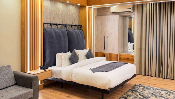 A modern hotel room with a wooden ceiling, a large bed with a headboard, a bedside table with a phone, a small sofa, a wooden floor, and a patterned rug.