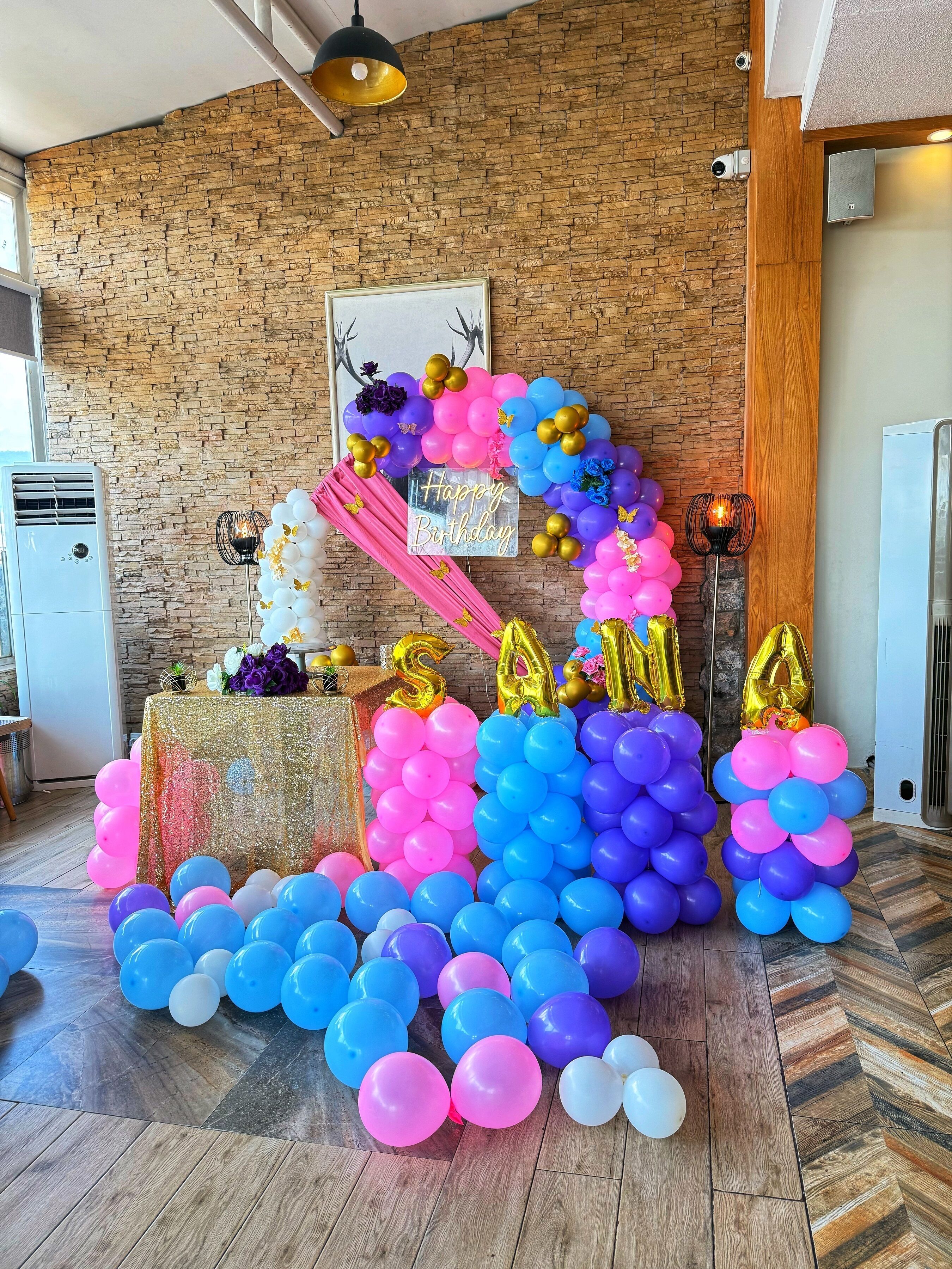 A room with a brick wall, a framed picture, and a birthday decoration with balloons.