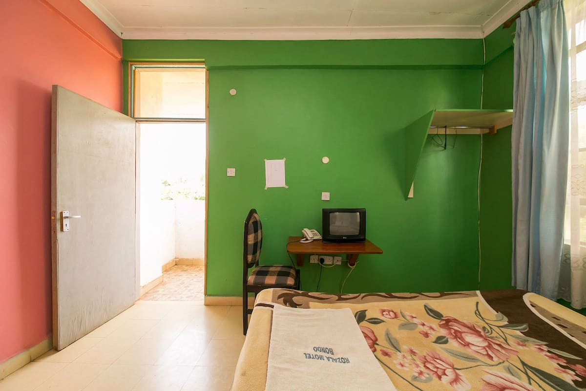 A room with a green wall, a bed with a floral bedspread, a chair, a small table with a television, and a window with curtains.