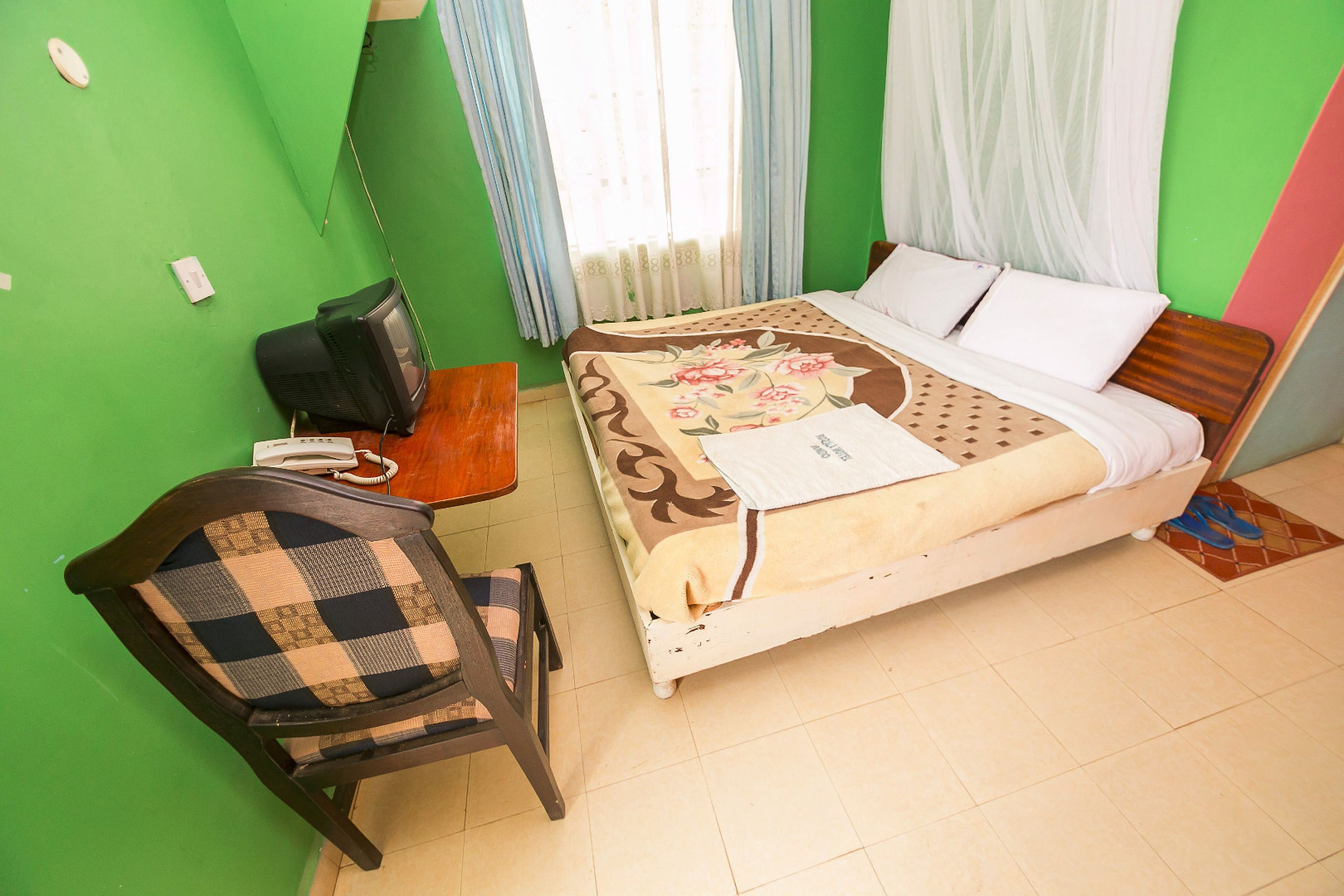 Una habitación con cama, una silla, escritorio con teléfono y ventana con cortinas.