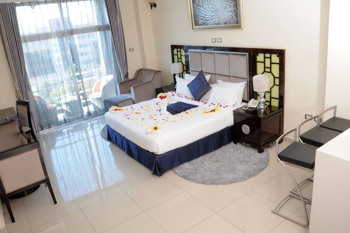 A hotel room with a large bed, a desk, a chair, and a view of the city through large windows.