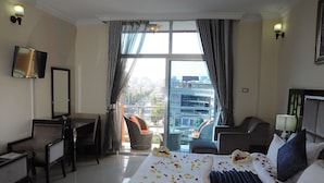 Quarto de hotel com cama, escrivaninha, cadeira e vista da cidade através de grandes janelas.