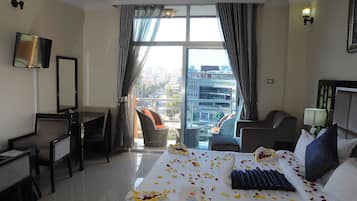 A hotel room with a bed, a desk, a chair, and a view of the city through large windows.