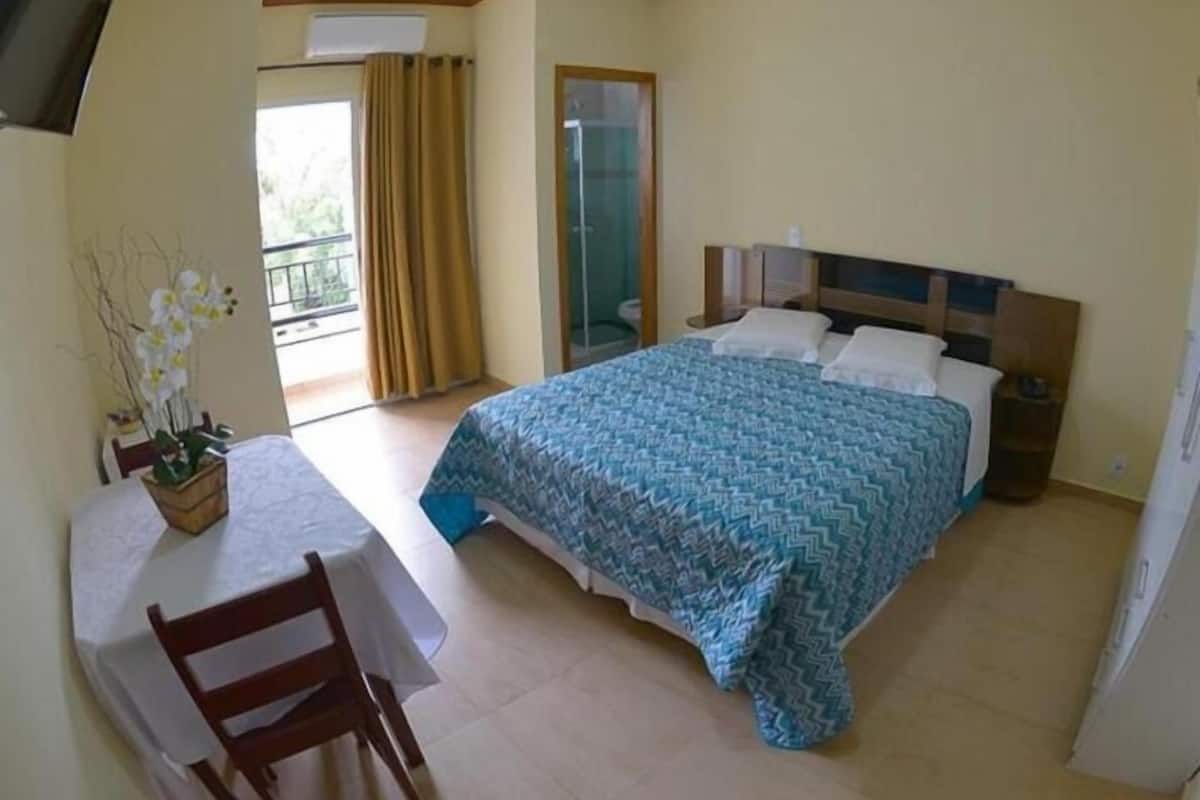 A hotel room with a bed, a table with a chair, a balcony with curtains, and a window.