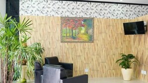 Un área de recepción con un muro de madera, un cuadro de un sendero bordeado de árboles, un televisor de pantalla plana y varias plantas en macetas.
