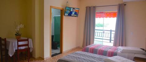 Habitación de hotel con dos camas, una mesa de comedor con sillas, televisor en la pared y un balcón con cortinas.