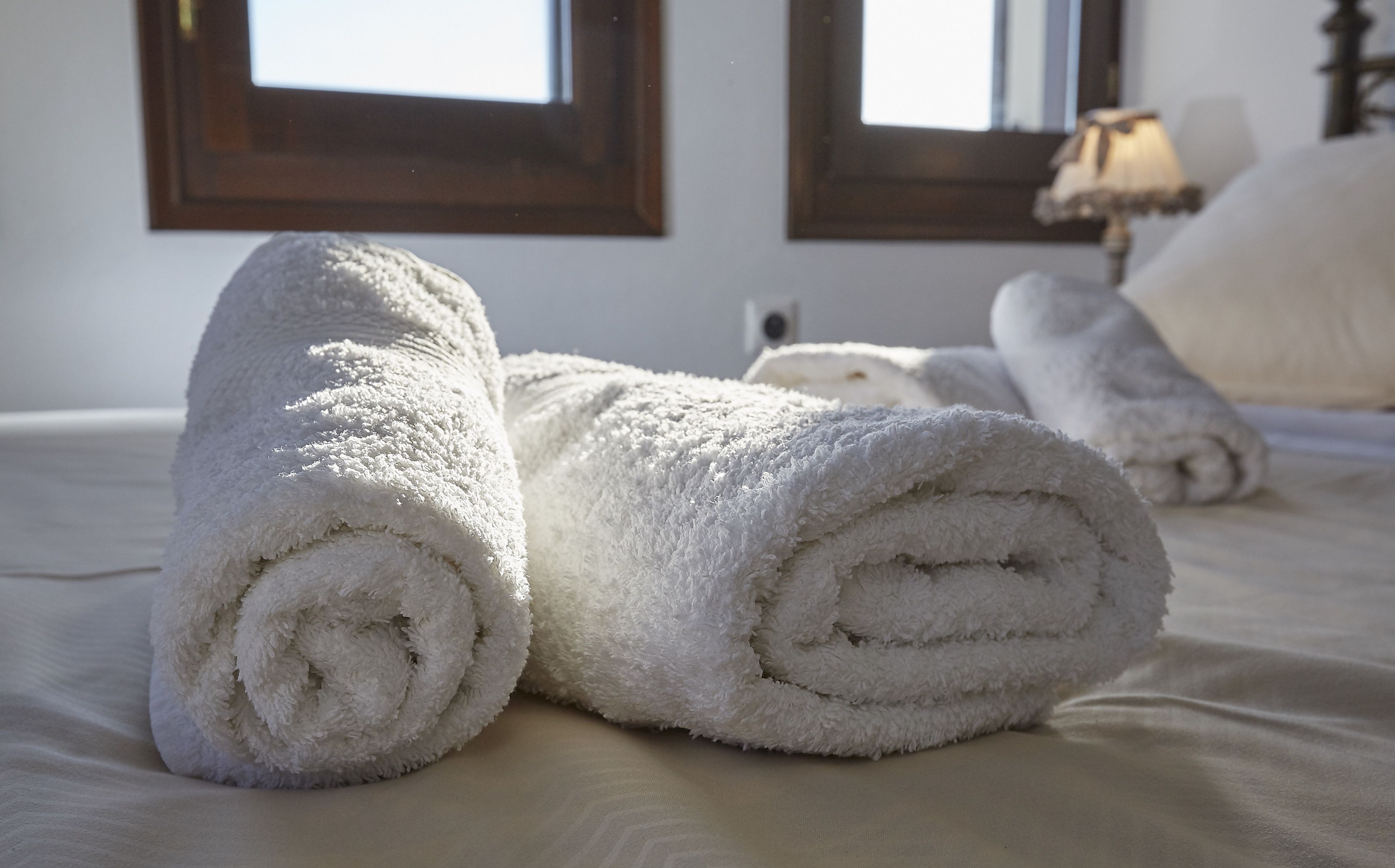 Neatly rolled white towels on a bed in a room with wooden headboards and a lamp.
