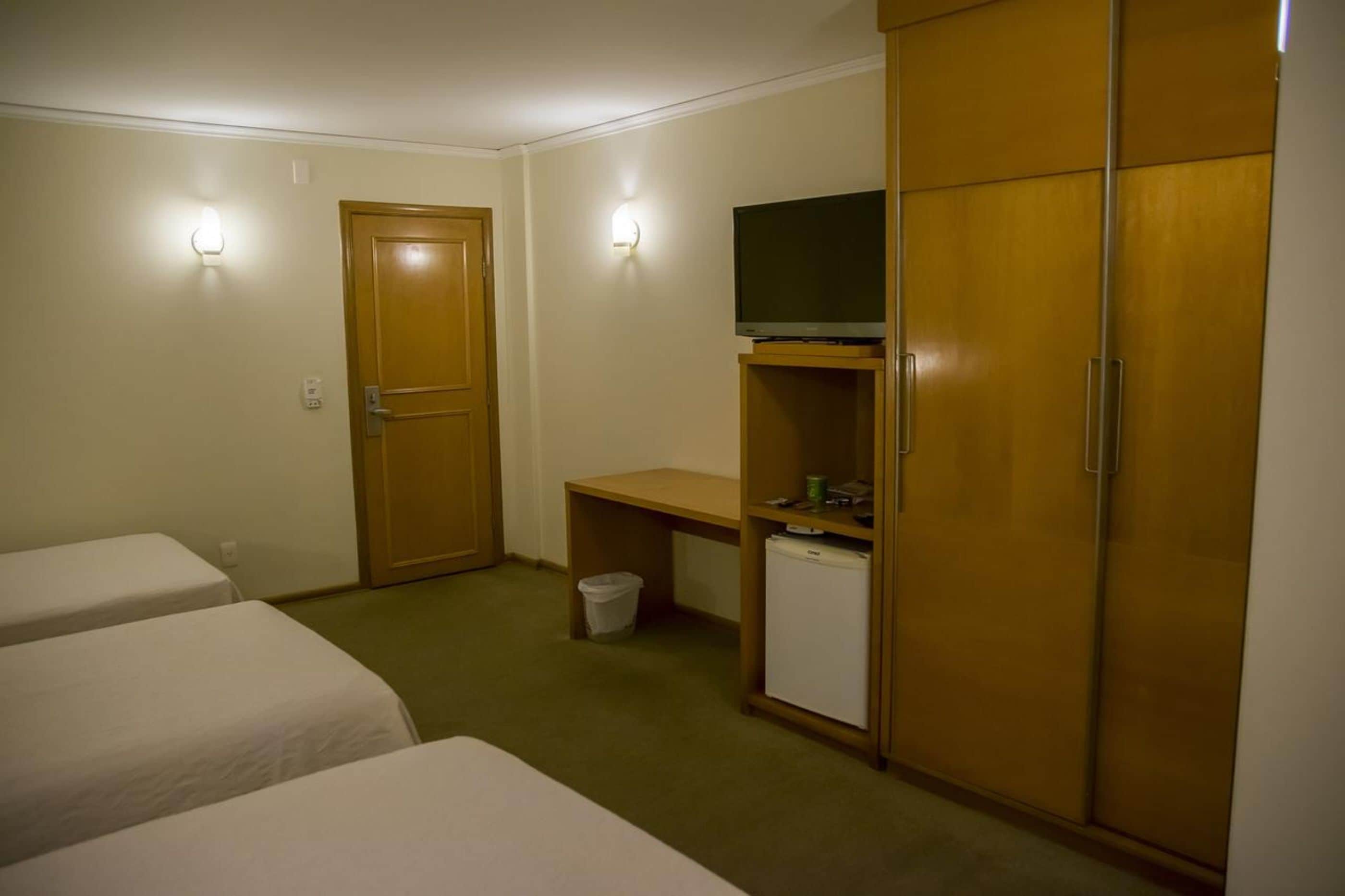 Une chambre d’hôtel avec deux lits simples, un bureau avec une télévision, une armoire et un petit réfrigérateur.