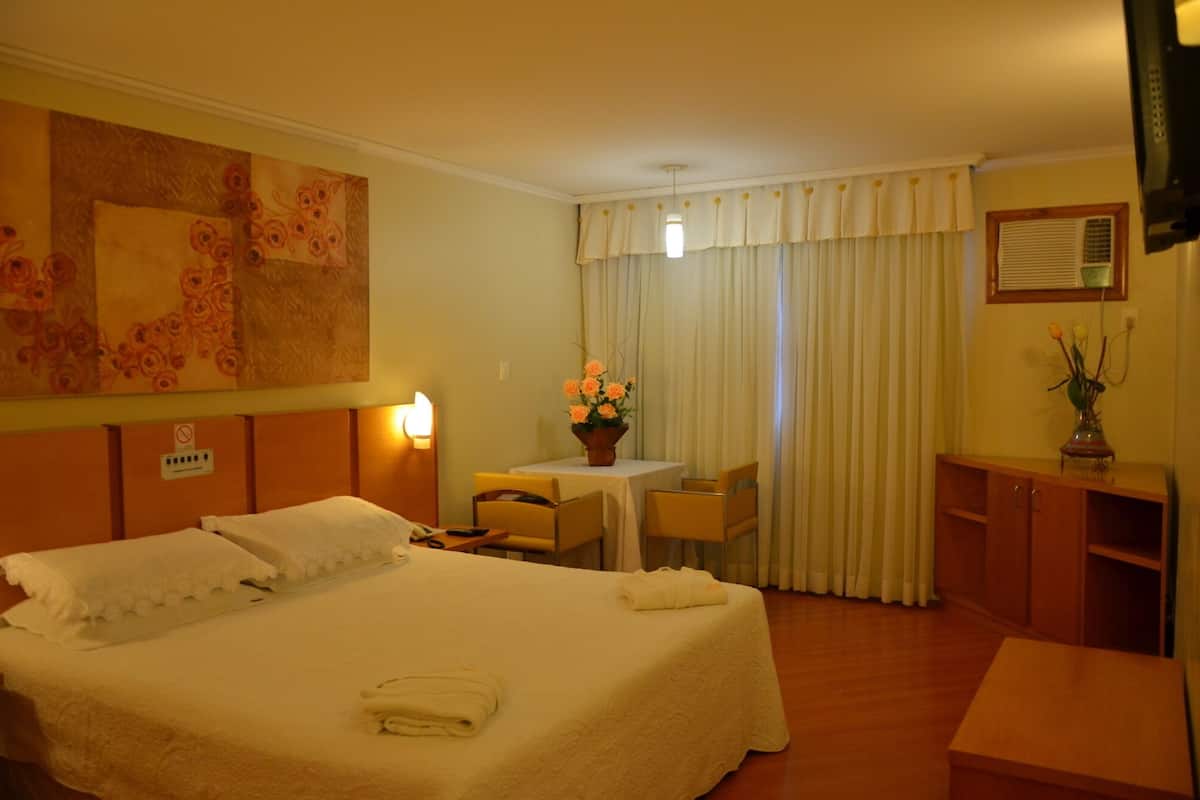 Ein Hotelzimmer mit einem großen Bett, einem Schreibtisch mit Stuhl, einem kleinen Tisch mit Blumenvase und einem an der Wand befestigten Fernseher.