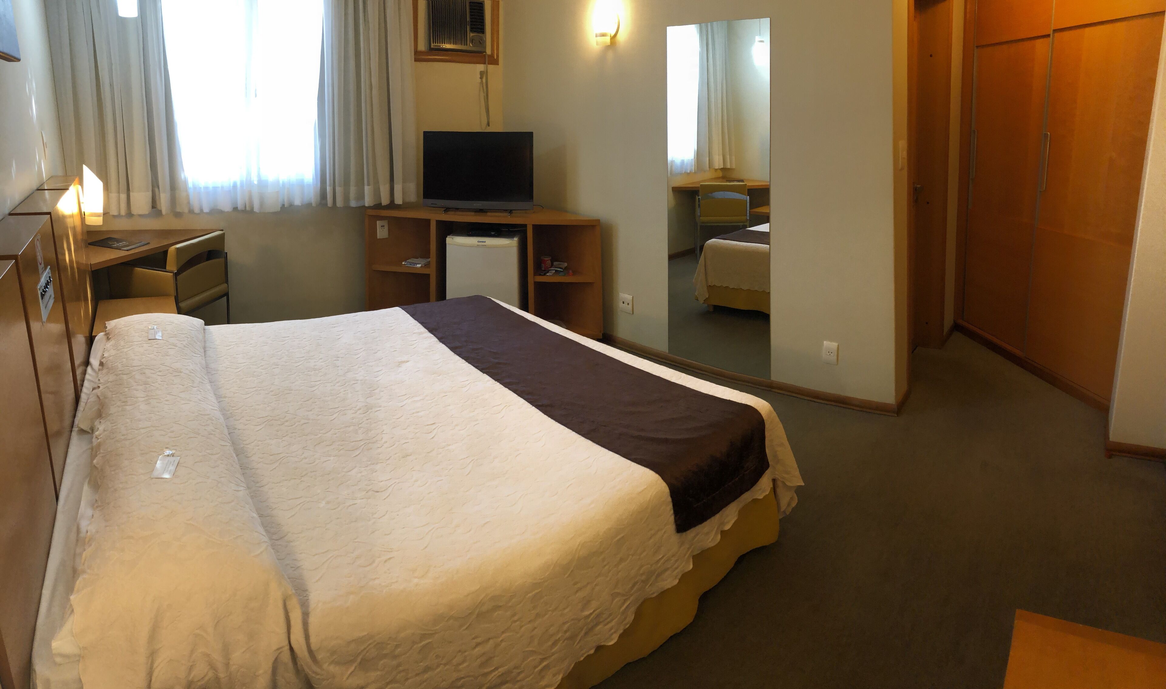 A hotel room with a bed, a desk with a lamp, a television, a wardrobe, and a mirror.