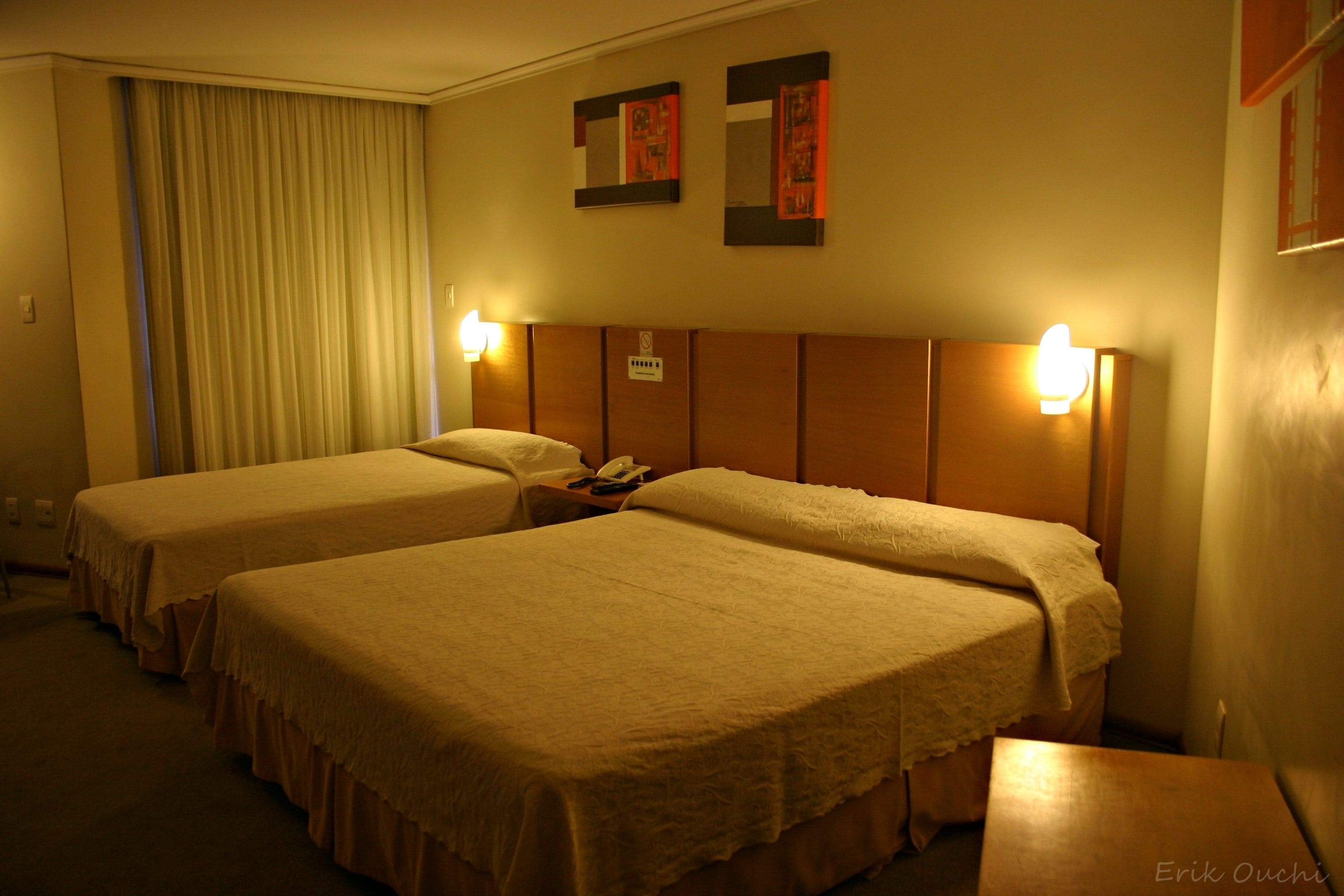 Habitación de hotel con dos camas individuales, una mesita de noche con teléfono e iluminación en la pared.
