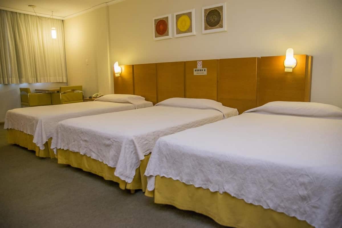 Ein Hotelzimmer mit drei Einzelbetten, jeweils mit weißer Bettdecke und gelbem, anliegendem Bettrunden. Zwei Wandleuchten und ein kleiner Tisch mit Stuhl befinden sich links.