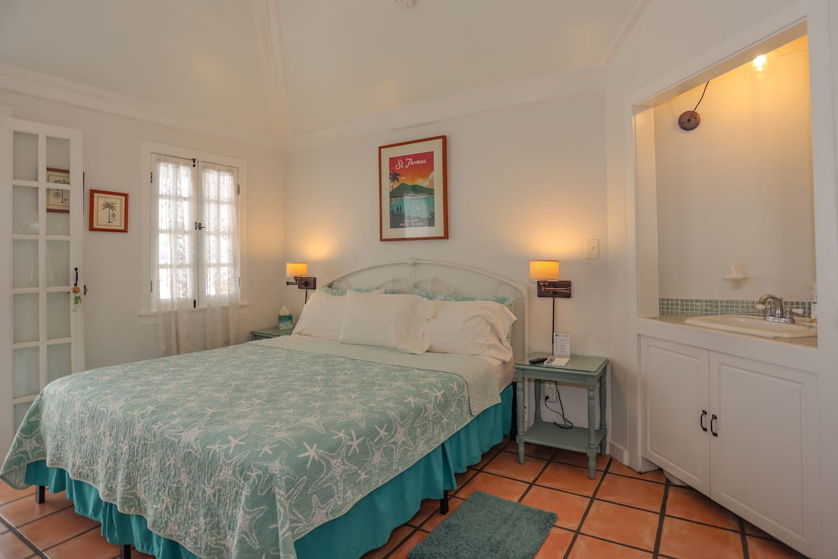 Ein Schlafzimmer mit Bett, Nachttischen, Fenster mit Vorhängen, einer Tür, einem gerahmten Bild an der Wand und einem Badezimmer mit Waschbecken und Spiegel.