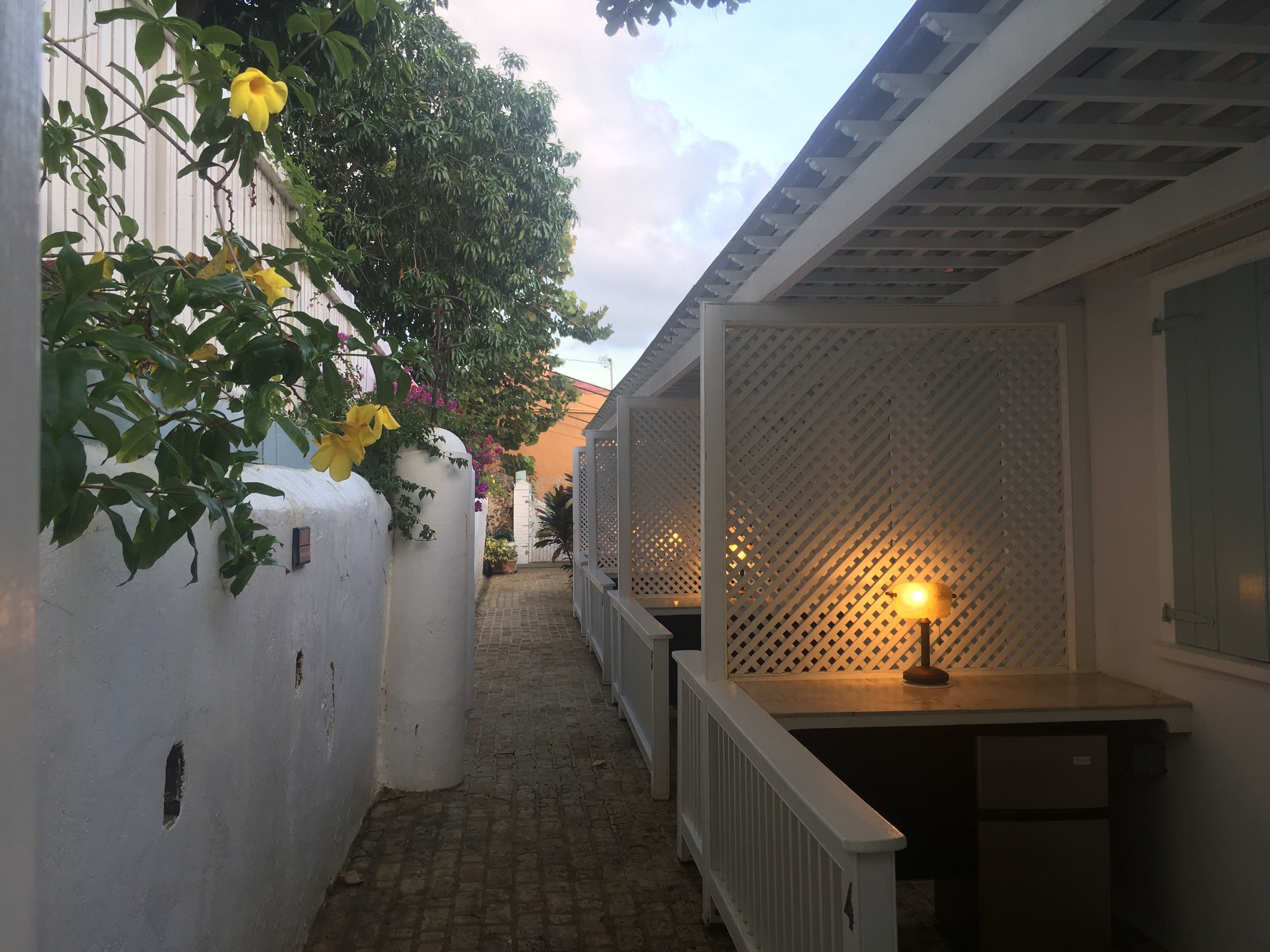 A narrow outdoor corridor with a white fence, a wooden bench, and a table with a lamp.