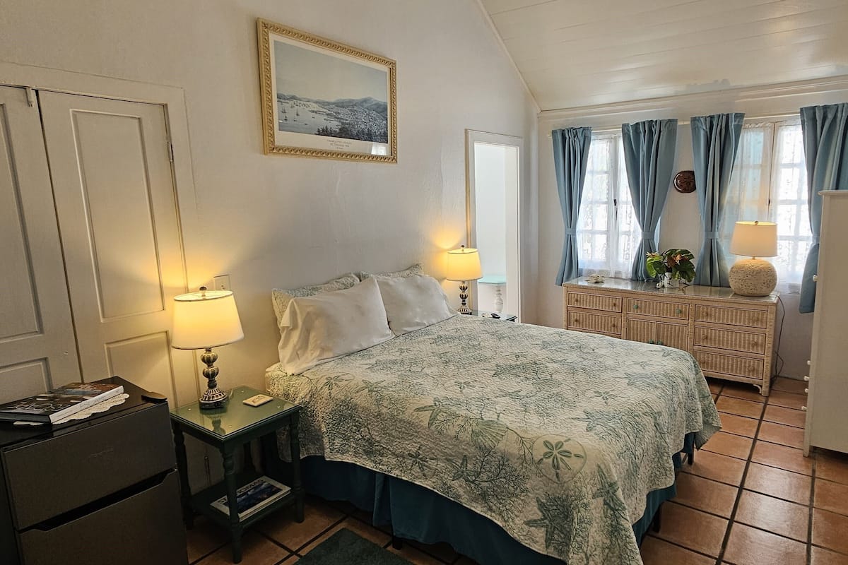 Ein Schlafzimmer mit einem Bett, Nachttischlampen, einem Schminktisch, einem Kühlschrank und einem Fenster mit Vorhängen.