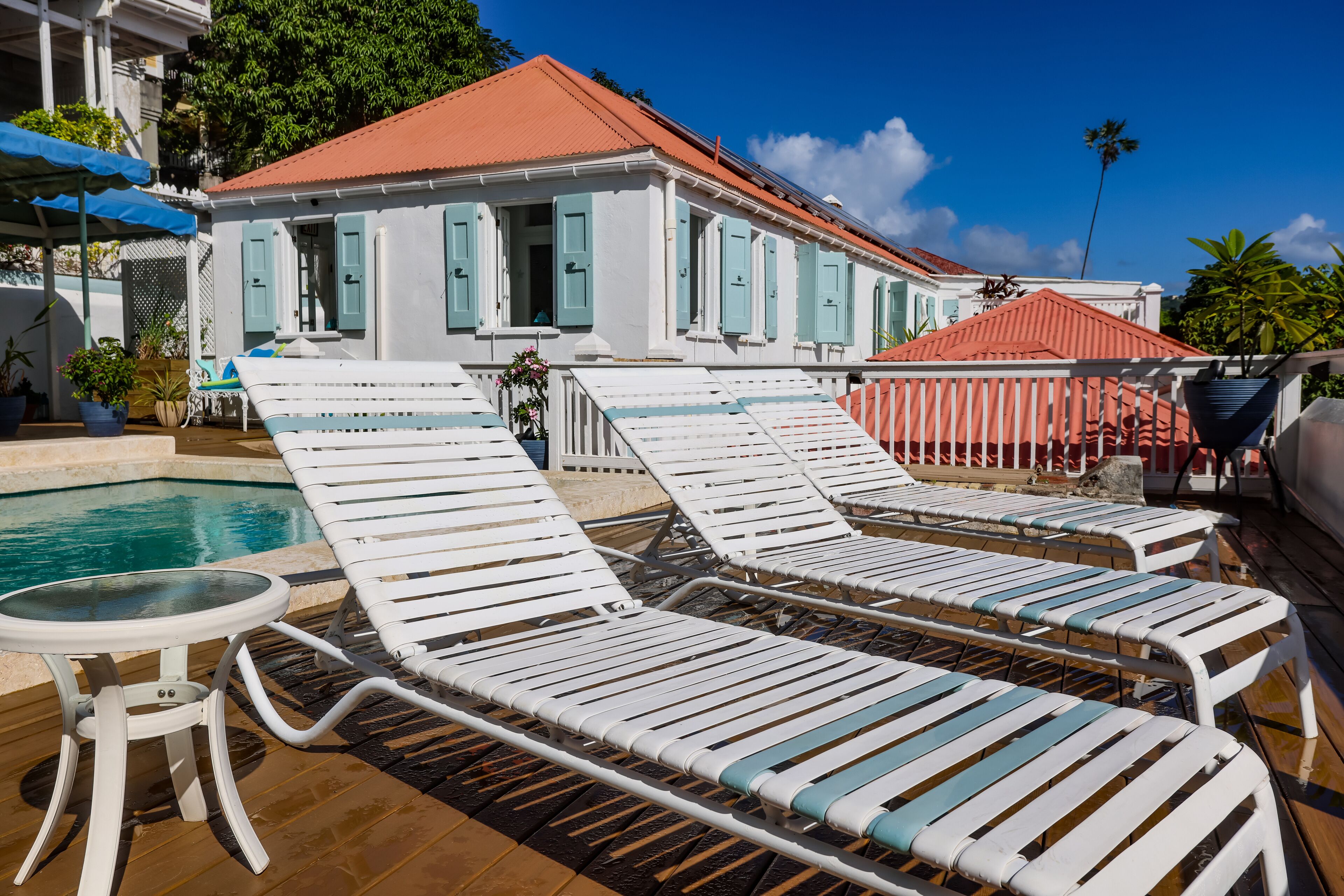 A poolside area with white lounge chairs and a small table, a building with a red roof, and a clear blue sky.