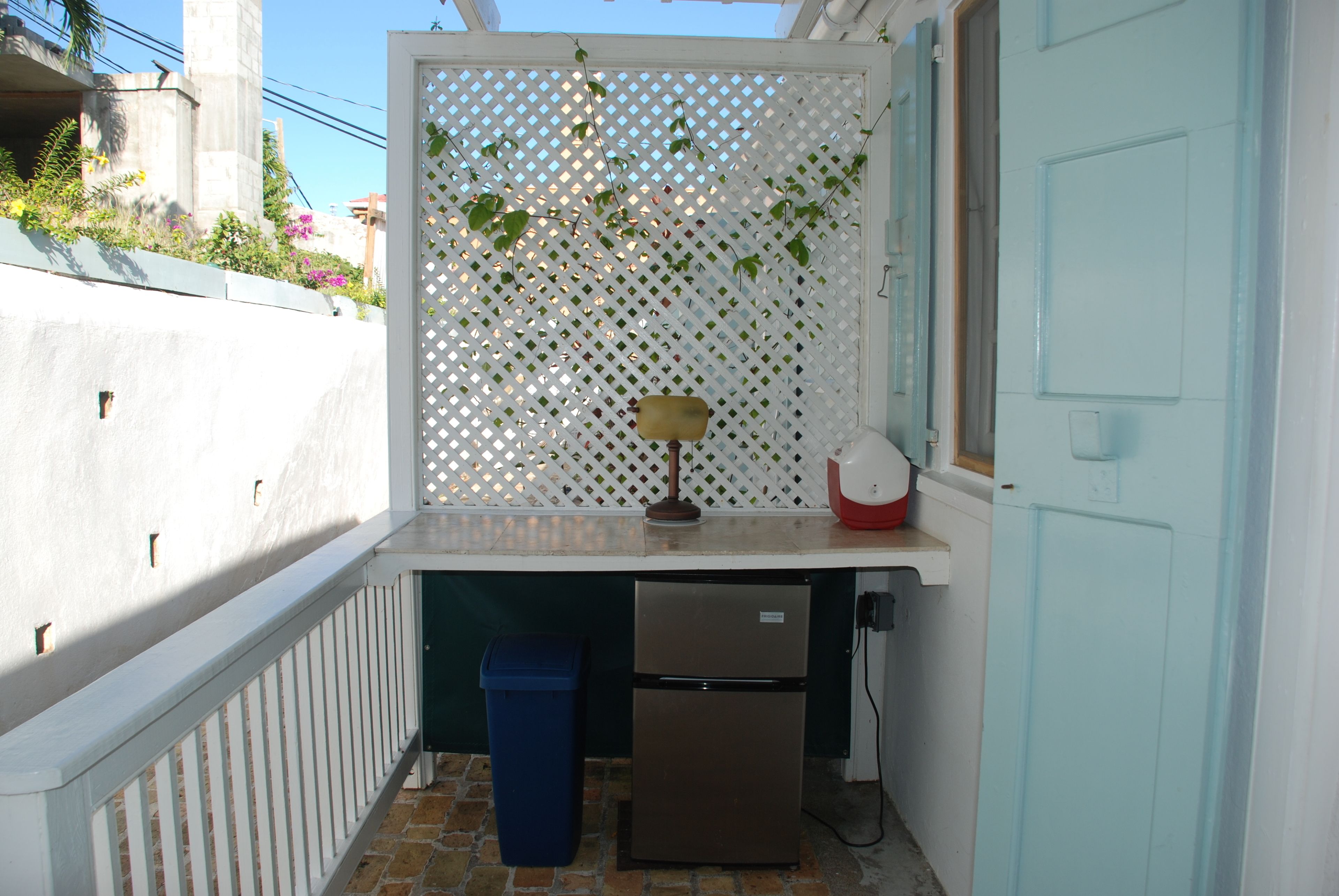 A compact kitchen with a refrigerator, a trash bin, and a small table with a lamp.
