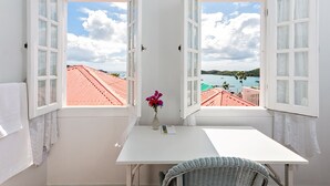 A room with a white table, a wicker chair, and a view of red-roofed buildings and a body of water through open windows. - At Home in the Tropics (St. Thomas)
