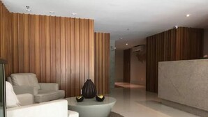 A modern lobby with a white sofa, a round coffee table with decorative items, and a reception desk. - London Residencial Design (Niteroi)